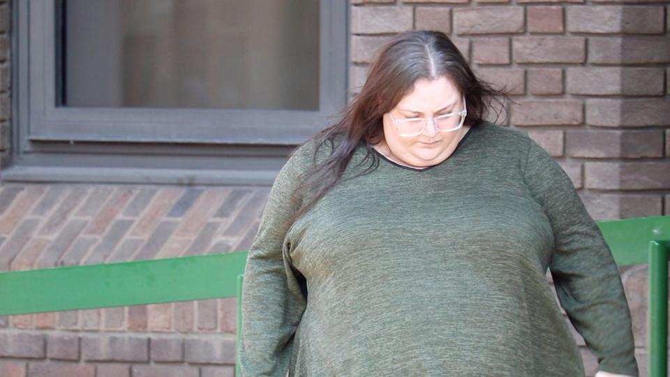 A woman wearing a dark green jumper and black leggings. She has straight brown hair and is wearing clear-framed glasses. She has her head down and wheeling a suitcase along with her